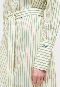 Χέρι γυναίκας κοντά στη μέση, που φοράει κρεμ πουκαμίσ dress με κάθετες πράσινες ρίγες και ταιριαστή ζώνη, μανσέτα του μανικιού κεντημένη με πράσινο λογότυπο-κείμενο.