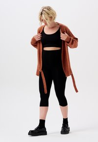 Rostfarbener gestrickter Cardigan mit langen Ärmeln und offener Vorderseite, über einem schwarzen gerippten Croptop und schwarzen Leggings, kombiniert mit schwarzen Plateau-Schuhen.