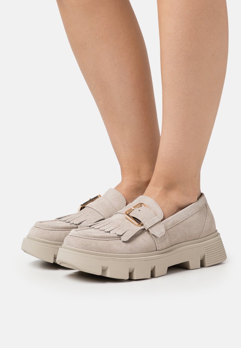 Mocassins en daim beige avec boucle dorée et détail à franges portés aux pieds, dotés de semelles en caoutchouc épaisses et rainurées sur fond blanc.