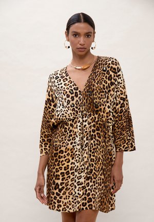 Mujer de cabello oscuro, con pendientes de aro dorados y vestido de escote en V con estampado de leopardo y cinturón de lazo, posando frente a un fondo liso y claro.