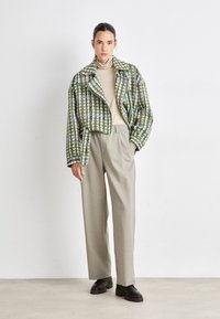 Veste cropped à carreaux verts et violets avec un large col, associée à un pantalon taille haute beige et des chaussures foncées sur un sol texturé.