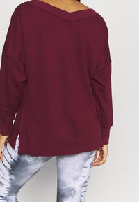 Bordeauxrotes Oversize-Sweatshirt mit weitem Ausschnitt, langen Ärmeln und Seitenschlitzen, kombiniert mit grauen Batik-Leggings. Weiche Stoffstruktur.