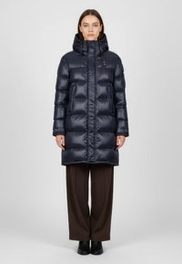 Námořnická péřová bunda s kapucí, prošívaná textura, zipové zapínání a nastavitelné šňůrky, v kombinaci s hnědými kalhotami s širokými nohavicemi a botami.