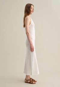Abito maxi bianco lavorato a maglia con corpetto aderente e orlo svasato. Presenta spalline sottili e un motivo verticale. Abbinato a sandali marroni con cinturini.