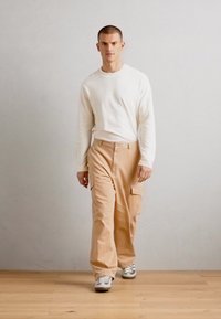 Beige cargo pants with side pockets, paired with a white long-sleeve shirt. Model walks on wooden flooring against a light gray wall.