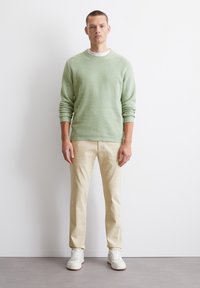 Hellegrüner Strickpullover mit gerippten Bündchen, kombiniert mit cremefarbenen Chinos und weißen Sneakers. Einfaches Design und lockere Passform, vor einem weißen Hintergrund stehend.