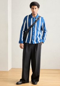 Niebiesko-biała koszula w paski z zapięciem na guziki, zestawiona z luźnymi czarnymi spodniami i czarnymi butami, noszona z torbą na ramię.