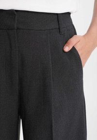 Mörkgrå byxor tillverkade av strukturerat tyg, med slät framsida, en veck och sidofickor. Midjan är slät och åtsittande.