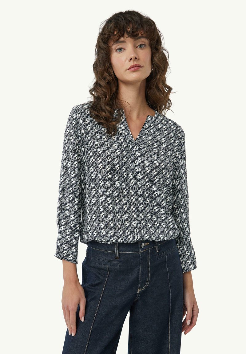 Langärmlige Bluse in Marineblau mit einem geometrischen weißen Muster, mit einem eingeschnittenen Ausschnitt und weichem Stoff; kombiniert mit dunkelblauen Jeans.