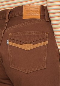Vaqueros de mezclilla marrones con un pecho de pana marrón y dos bolsillos traseros. La etiqueta presenta texto en relieve y detalles de diseño en la cinturilla.