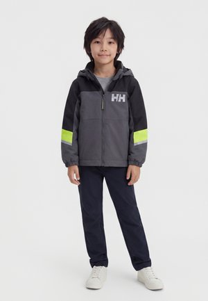 Jeune garçon debout portant une veste zippée noire et grise avec des bandes jaune fluo et un pantalon foncé avec des baskets blanches sur fond blanc.
