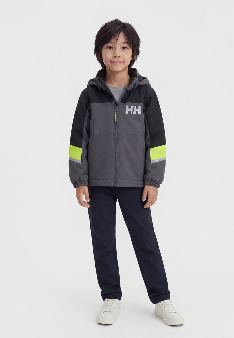 Jeune garçon debout portant une veste zippée noire et grise avec des bandes jaune fluo et un pantalon foncé avec des baskets blanches sur fond blanc.