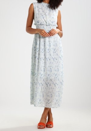 Vrouw draagt een mouwloze, lichtblauwe plissé midi-jurk met bloemenprint, gecombineerd met oranje open-toe sandalen met hak, tegen een witte achtergrond.