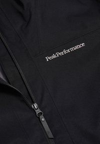 Schwarze Jacke aus strapazierfähigem Stoff mit Reißverschluss und weißem "PeakPerformance"-Logo als Stickerei auf der Vorderseite.