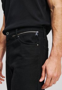 Vaqueros de mezclilla negra con acabado texturizado, que cuentan con un bolsillo frontal con cremallera, bolsillos estándar y detalles en hardware plateado.