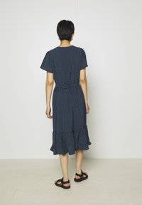 Osoba s krátkými tmavými vlasy, oblečená v námořnické modré šatech s bílými puntíky a černými sandály, stojí čelem ke bílé zdi.