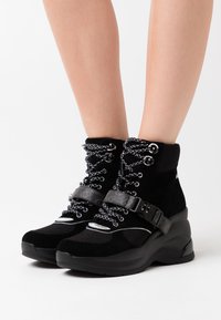 Czarne sneakersy typu high-top wykonane z zamszu i siatki, z metalowymi dodatkami w kolorze srebrnym, teksturowanymi sznurowadłami oraz podniesioną podeszwą dla zwiększenia wysokości.