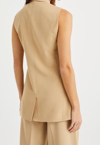 Ärmlös beige topp med skräddarsydd design, synliga sömmar, bakslits och mjukt, lätt material. Passar bra för lager-på-lager eller som ensam plagg.