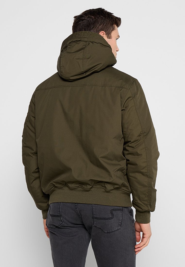 Veste à capuche vert olive avec une finition lisse et mate, des poignets et un ourlet côtelés, et une coupe décontractée. Pas de marques ou d'ornements visibles.