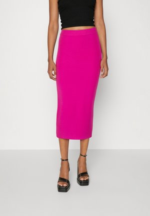 Kvinde iført en knælang, tætsiddende nederdel i klar pink, sort cropped ærmeløs top og sorte platformsandaler med remme om anklerne.