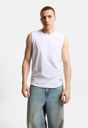 Młody mężczyzna z krótkimi włosami, ubrany w białą koszulkę bez rękawów i luźne jasnoniebieskie dżinsy, stoi na jednolitym białym tle.