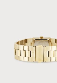 Χρυσό μεταλλικό ρολόι με τετράγωνη πρόσοψη, σχεδίαση αλυσίδας για το μπράτσο, γυαλιστερό φινίρισμα και μια μικρή κορώνα στο πλάι για ρυθμίσεις.