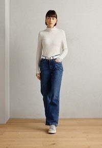 Maglietta bianca a maniche lunghe con colletto alto, dal design aderente, abbinata a jeans blu di denim larghi e sneaker grigio chiaro. Accento con cintura semplice.