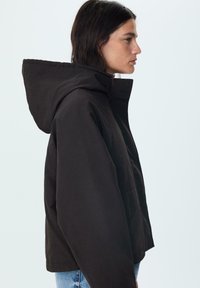 Čierna kapucňová bunda so striktným dizajnom, s prednou vreckom a textúrovaným povrchom. Silueta je moderná a široká.