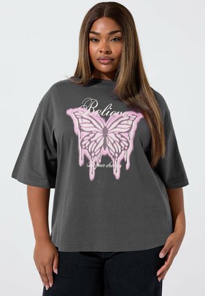 Donna con capelli lunghi e lisci che indossa una maglietta larga grigio scuro con una farfalla rosa e la scritta "Credi nei tuoi sogni."