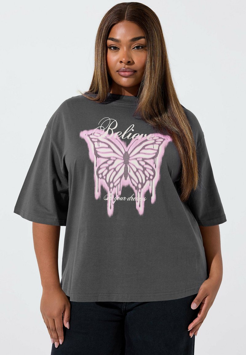 Kobieta o długich, prostych włosach, ubrana w luźny, ciemnoszary T-shirt z różowym motylem i napisem "Uwierz w swoje marzenia."