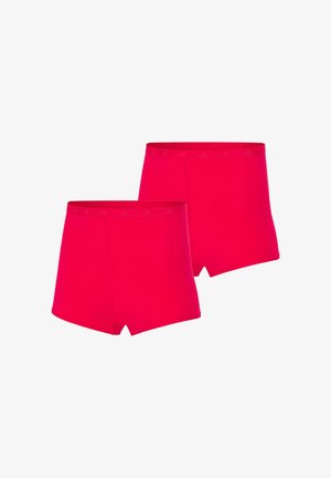 Kobieta stojąca boso, ubrana w dopasowany, jaskrawoczerwony biustonosz i szorty z wysokim stanem na jednolitym białym tle.