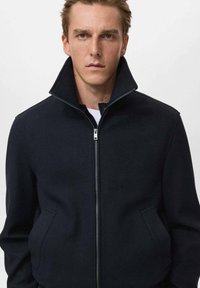 Giacca in misto lana blu navy con colletto alto, zip frontale e tasche laterali. Texture liscia con silhouette strutturata.