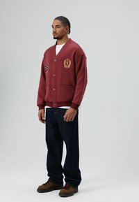 Bordeauxrode cardigan met V-hals, knopen aan de voorkant, twee zakken, geborduurd logo en cijfers op de borst. Gecombineerd met donkere jeans en bruine laarzen.