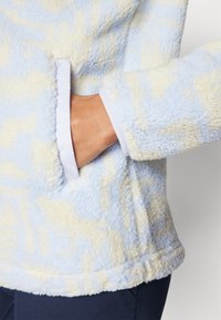 Pullover in pile di colore blu chiaro e crema. Presenta una finitura morbida e testurizzata, maniche lunghe e una tasca laterale con bordatura bianca.