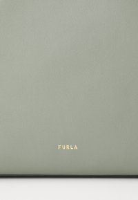 Zelena teksturirana usnjena površina z embosiranim Furla logotipom v zlati barvi na dnu. Površina se zdi gladka z rahlo zrnato obdelavo.