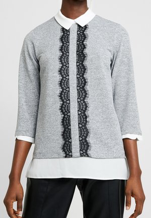 Vrouw draagt een grijze top met lange mouwen, witte kraag en manchetten, met een verticale zwarte kanten streep aan de voorkant, gecombineerd met zwarte broek.