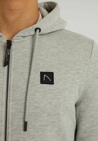 Acercamiento de una persona que lleva puesta una sudadera con cremallera de color gris claro, con un parche de logo cuadrado negro y cordones en la zona del pecho.