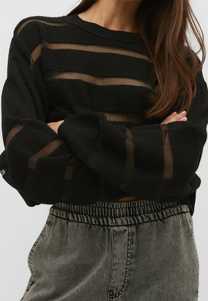 Femme portant un pull noir avec des rayures horizontales transparentes et des manches amples, associé à un jean gris délavé taille haute avec une ceinture élastique.