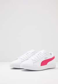 Para białych sneakersów Puma z różowym paskiem po boku, zawiązanymi sznurówkami, umieszczona na białej powierzchni na tle gładkiej białej ściany.