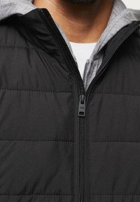 Schwarze Steppweste mit Reißverschluss, ausgestattet mit einer grauen Kapuzenfütterung und diagonaler Steppmusterstruktur. Leichtes und isoliertes Design.