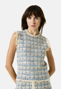 Vrouw met donker haar in een mouwloze blauwe en beige tweed top en bijpassende rok, staand met handen achter de rug op een witte achtergrond.