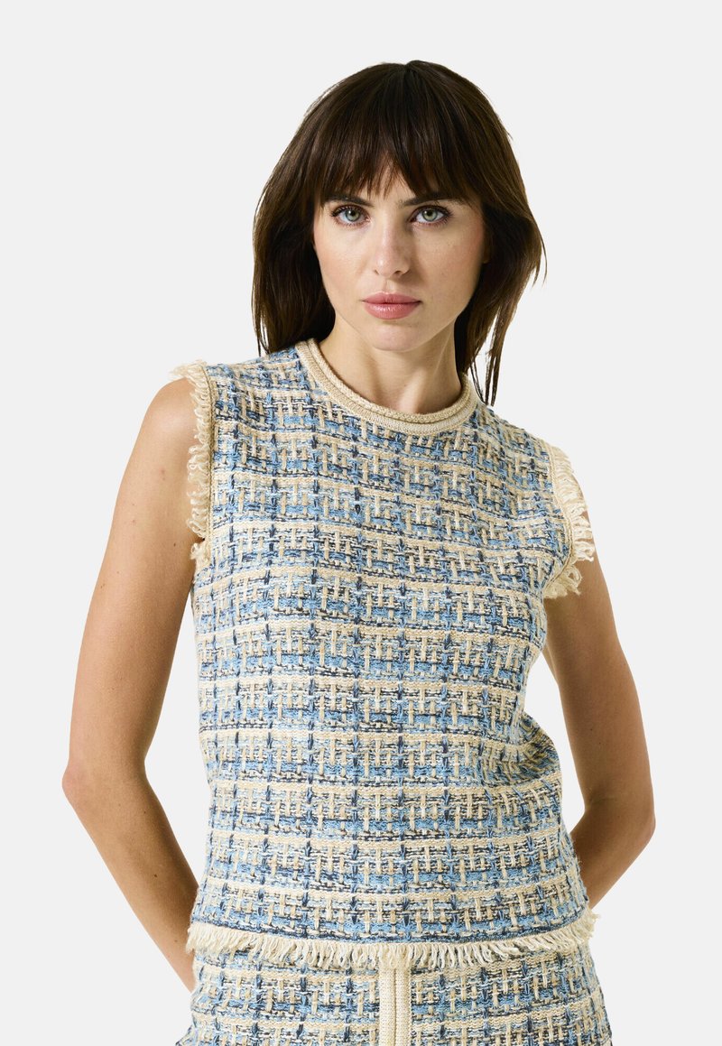 Vrouw met donker haar in een mouwloze blauwe en beige tweed top en bijpassende rok, staand met handen achter de rug op een witte achtergrond.
