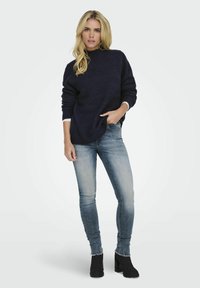 Sötétkék kötött pulóver magas garbóval, világos koptatott skinny farmerral és fekete bokacsizmával párosítva. Simább textúra bordás részletekkel.
