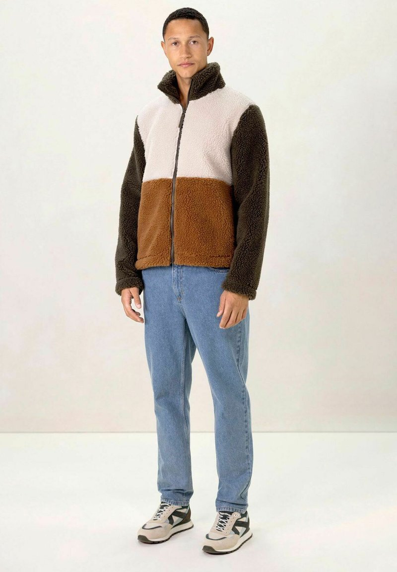 Veste en polaire avec col montant, présentant un haut beige, des manches marron et une partie inférieure marron. Portée avec un jean bleu clair et des baskets.