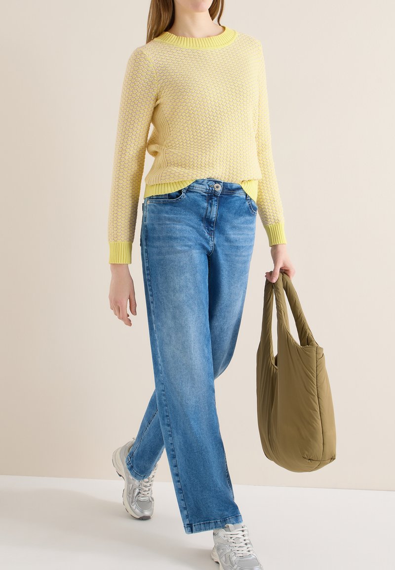 Femme portant un pull jaune texturé, un jean bleu, des baskets argentées, tenant un sac fourre-tout matelassé vert olive en marchant.