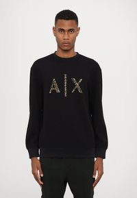 Sudadera negra hecha de tela suave, decorada con un logo texturizado dorado "AIX" en la parte frontal. Mangas largas y un escote redondo con puños acanalados.