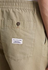 Beige cotton shorts with an elastic waistband, back pocket featuring a brown button, and a fabric tag displaying brand and text.