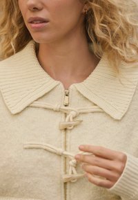 Cream-colored sweater with a ribbed collar, zip closure, decorative toggles, and textured fabric. Features a soft, inviting appearance.