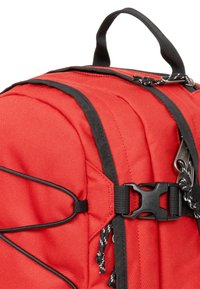 Zaino rosso con accenti neri, caratterizzato da un tessuto testurizzato, spallacci regolabili e una chiusura anteriore con fibbia e dettagli in cordino elastico.