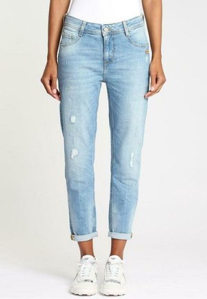 Jasnoniebieskie slim fit jeansy podwinięte z lekkim przetarciem, noszone z białymi masywnymi sneakersami na stojącej osobie.
